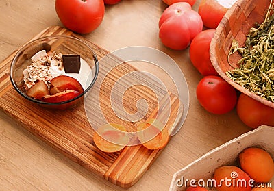 A smoothie bowl with apricots placed on a wooden table. There are tomatoes and herbs around. Stock Photo