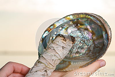 Smudging Ritual using burning thick leafy bundle of White Sage in bright polished Rainbow Abalone Shell on the beach at sunrise Stock Photo