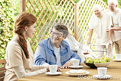 Snack time with the elderly on the patio of a nursing home. Volunteer lady talking to a senior woman sitting by a table. Two men Stock Photo