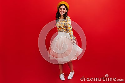 Snapshot full-length of curly dark-haired girl in bright outfit. Woman in beret and fluffy skirt is spinning on red Stock Photo