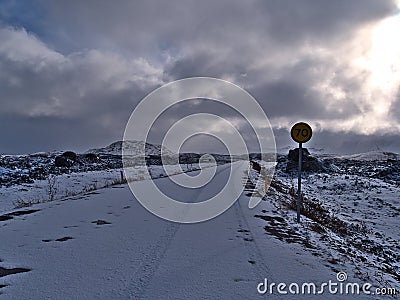 Snow-covered country road with tire marks and speed limit sign leading through lava field with rocks near HafnarfjÃ¶rÃ°ur. Stock Photo