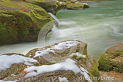 Snow on mossy rocks beside smooth cool flowing river water Stock Photo