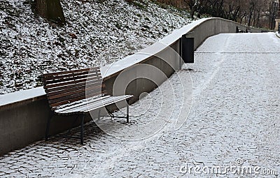 Snowy benches near the supporting concrete gray wall in the park. Paving and metal low fences protect ornamental flower beds from Stock Photo