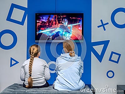 Sochi, Russia - 13 October 2019. Two girls sit on a gray pouf and look into the TV screen hanging on a gray blue wall Editorial Stock Photo