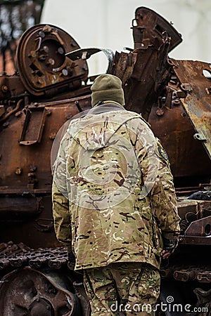 Soldier in uniform near a burnt and melted rusty wreckage of a Soviet Russian-made tank Editorial Stock Photo