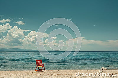 A solitary red chair faces the tranquil blue sea, offering a peaceful retreat under the vast, cloud filled sky Stock Photo