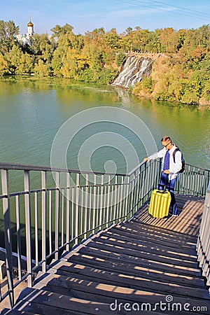 Solo traveling man with backpack and yellow luggage on the wooden bridge with church and waterfall background Stock Photo