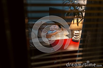 Someone is peeping through the blinds on window at woman lying in bed in her bedroom. Concept of voyeurism and criminal Stock Photo