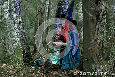 The sorceress in a hat and a black cloak in the forest charms over a luminous crystal ball. Horizontal photo Stock Photo