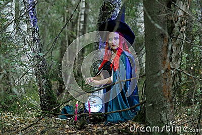 The sorceress in a hat and a black cloak in the forest charms over a luminous crystal ball. Horizontal photo Stock Photo