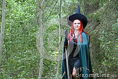 A sorceress with red hair in a pointed hat and a black cloak in the forest is engaged in charms with a candle. Stock Photo