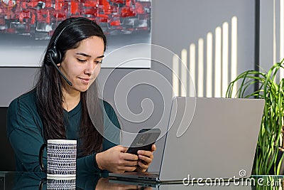 South Asian woman having a business call in headphones at a desk, typing on a phone in home office Stock Photo