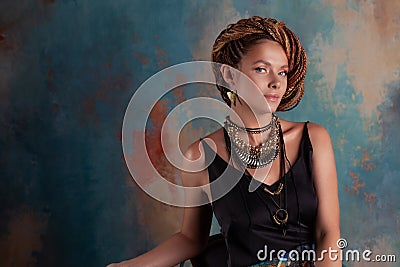 Southern spices. A beautiful young blue-eyed woman with afrocos and large ornaments against textured rustic style wall . Stock Photo