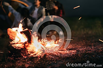 Sparking bonfire with tourist people sit around bright bonfire near camping tent in forest in summer night background. Group of Stock Photo