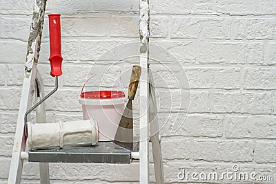 A spatula, bucket and textured roller under the brick stand on a metal stepladder on the background of a white wall. Stock Photo