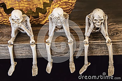 Speak no evil See no evil Hear no evil skeletons sitting on edge of a table with giant scarey metal pumpkin mouth behind them for Stock Photo