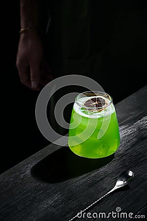Special green cocktail of mixology, lemon rum, garnished with dried fruit, on black bar table, bartender in the background Stock Photo