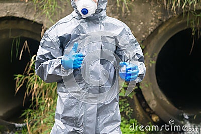 A specialist in protective clothing and a mask holds in one hand a flask with a reagent and shows the thumb up sign with the other Stock Photo