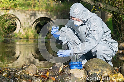 A specialist in a protective suit and respirator analyzes the water in the river near the existing plant, using a flask and a Stock Photo