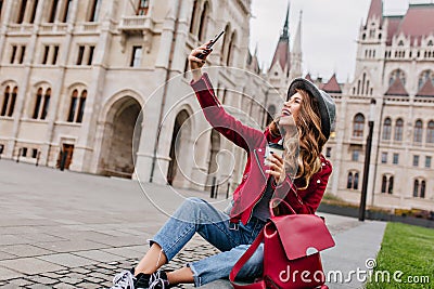 Spectacular young woman sitting on the ground with backpack and taking picture of herself. Outdoor shot of elegant Stock Photo