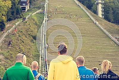 Spectators on sport event cheering for conmpetitors running upwards on ski jump. Editorial Stock Photo