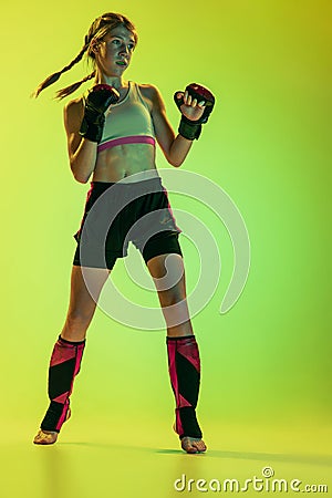 Dynamic portrait of athletic girl, MMA fighter training isolated on gradient yellow-green background in neon. Concept of Stock Photo