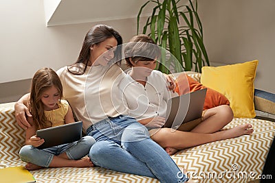 Spending time with kids. Young beautiful caucasian family, mother and her two cute little kids relaxing on sofa, using Stock Photo