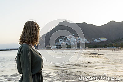 Spiritual adolescent in green dress admire the sunset and the sea town on a hill in Tenerife. Blonde romantic woman look the Stock Photo