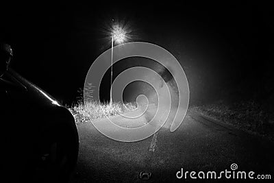 A spooky ghost of a woman in the road below a single street light in the countryside. Lit up by car headlights at night. With a Stock Photo