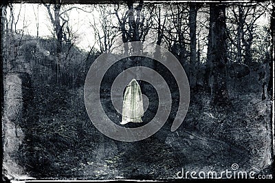 A spooky ghostly white figure standing on a path in a forest in winter. With a grunge, vintage black and white edit Stock Photo