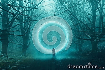 A spooky hooded figure, standing in front of a magical glowing portal floating in a forest. In a mysterious, foggy, winter forest Stock Photo