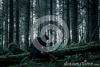 A spooky lone hooded figure sitting on a moss covered log in a forest in winter. With a muted edit Stock Photo