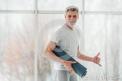 Sport its life. Senior man standing in the white gym holding yoga mat and showing gesture call me Stock Photo