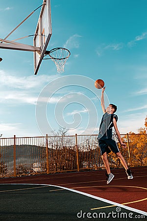 Sports and basketball. A young teenager in a blue tracksuit throws a jump ball into the basket. Blue sky. Vertical Stock Photo