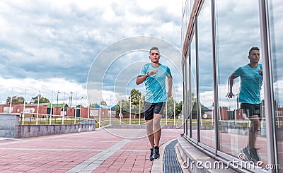 Sports man runs in jump workout, glass windows in background, runner in sportswear, a summer afternoon training in city Stock Photo