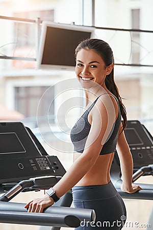 Sporty beauty. Cheerful and beautiful young woman in sportswear is standing on treadmill at gym and looking at camera Stock Photo