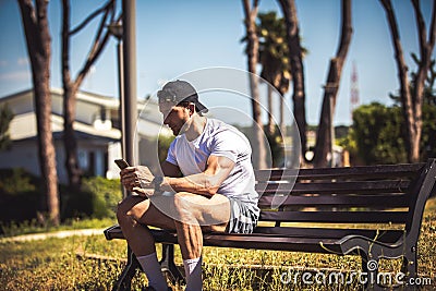 Sporty man personal trainer sitting looking at his phone on a bench in an outdoor park. Muscle body and well defined. Sporty Stock Photo