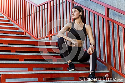 Sporty slim young beautiful woman in black fashionable sportwear sitting on the red stairs and relaxing during outdoor exercises Stock Photo