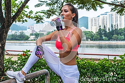 Sporty young female athlete taking a break after exercising or running, standing and drinking water from bottle in park Stock Photo