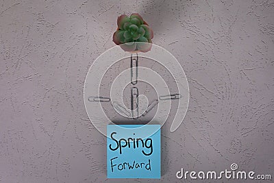 Spring forward sign handwritten on blue posted sticky note with small house desk plant and paper clips shape of flower Stock Photo