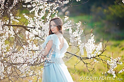 Spring touch. Happy beautiful young woman in blue dress enjoy fresh flowers and sun light in blossom park at sunset. Stock Photo