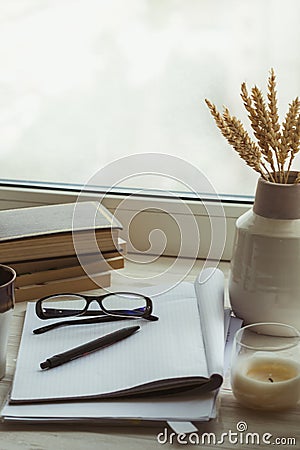 A stack of books, cup of coffee, bouquet of kernels, glasses, notebook and pen over a window. Stock Photo