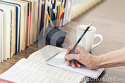 Stack of books education background, female hand makes notes in open notebook. Glasses, pens and pencils in holder, cup of tea Stock Photo