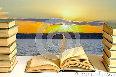 A stack of books and an open book against the backdrop of a sunset over the river. Reading visualization concept Stock Photo