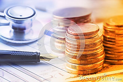 Stack of coins with a fountain pen and an chest-piece of a stethoscope on a table. Stock Photo