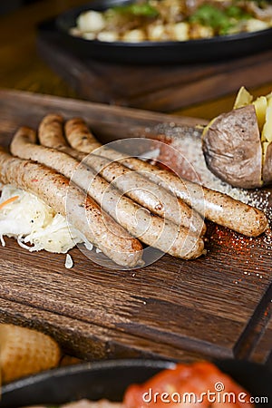 Stack of cooked sausages served on a rustic wooden board with sauce and baked potatoes. Stock Photo