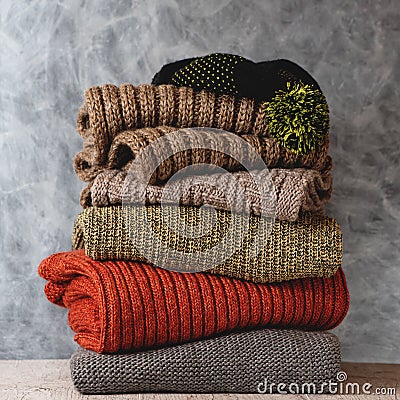 Stack of folded knitted wool women sweaters, scarf, hat with pompom on shelf against gray wall. Season of warm knitwear Stock Photo