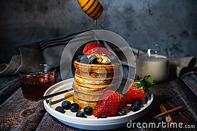 A stack of pancakes with pouring honey, blueberry and strawberry and whip cream on top in plate on wooden table Stock Photo