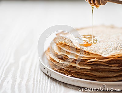 Stack Russian homemade thin pancakes on white plate and honey flows from wooden spoon. Traditional wheat pancakes for Shrovetide Stock Photo