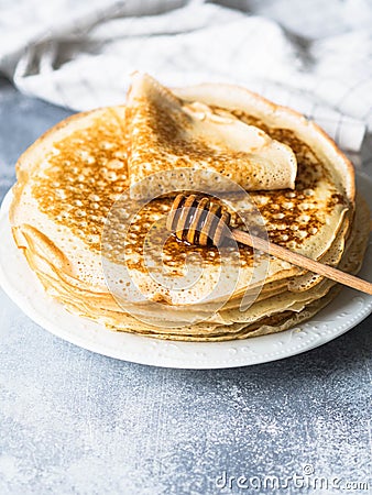 Stack Russian homemade yeast pancakes on white plate, wood honey spoon on grey table. Traditional wheat crepes for Shrovetide Stock Photo